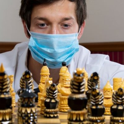 A close up of the face of a man wearing a surgical mask behind a chess board set with playing pieces